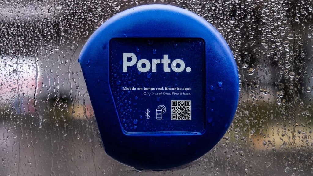 A Porto beacon behind rain-covered glass, carrying the city mark, bluetooth and NFC symbols and a QR code, above the words city in real time in Portuguese and English